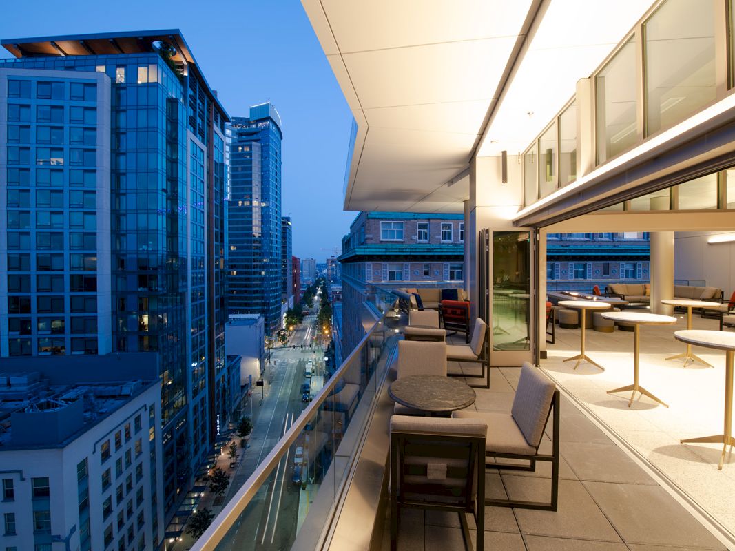 A modern outdoor balcony with tables and chairs overlooks a city street lined with tall buildings at twilight.