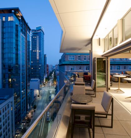 A modern rooftop balcony overlooking a cityscape at dusk, with seating areas and buildings illuminated by streetlights and interior lights.
