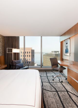 The image shows a modern hotel room with a large bed, desk, chair, TV, and a view of buildings and water through large windows.