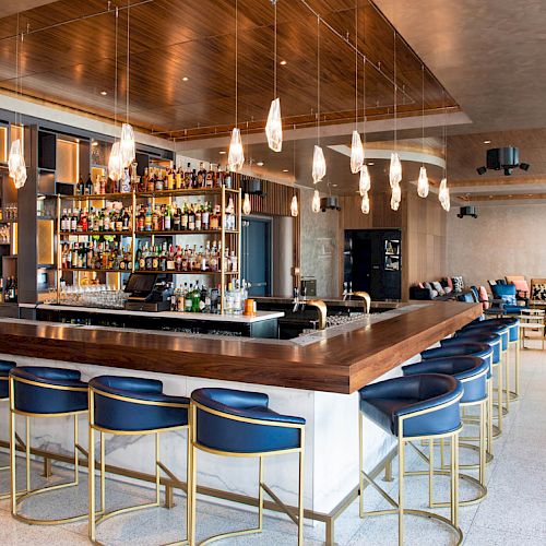 A modern, stylish bar with a sleek, well-stocked counter, elegant lighting, and seating for patrons. Cozy lounge area visible in the background.