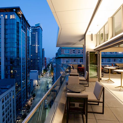 A rooftop terrace with modern furniture overlooks a cityscape at dusk, featuring tall buildings and a softly lit street.