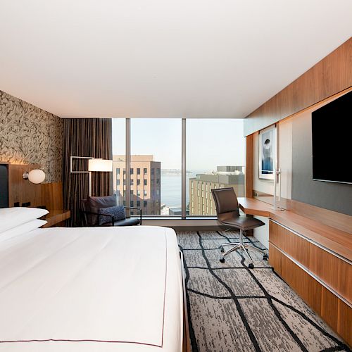 A modern hotel room features a large bed, wall-mounted TV, desk, and window with an urban view, accented by stylish decor and lighting.
