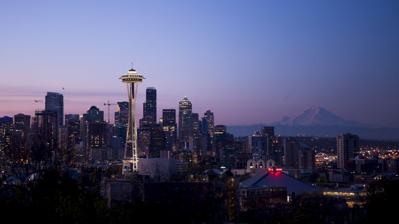 A city skyline at dusk with a prominent tower, surrounding buildings, and a mountain in the background under a purple sky.
