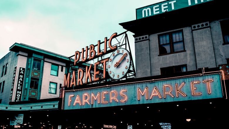 The image shows a historic public market with neon signs for "Public Market" and "Farmers Market" and a clock on the facade of a building.