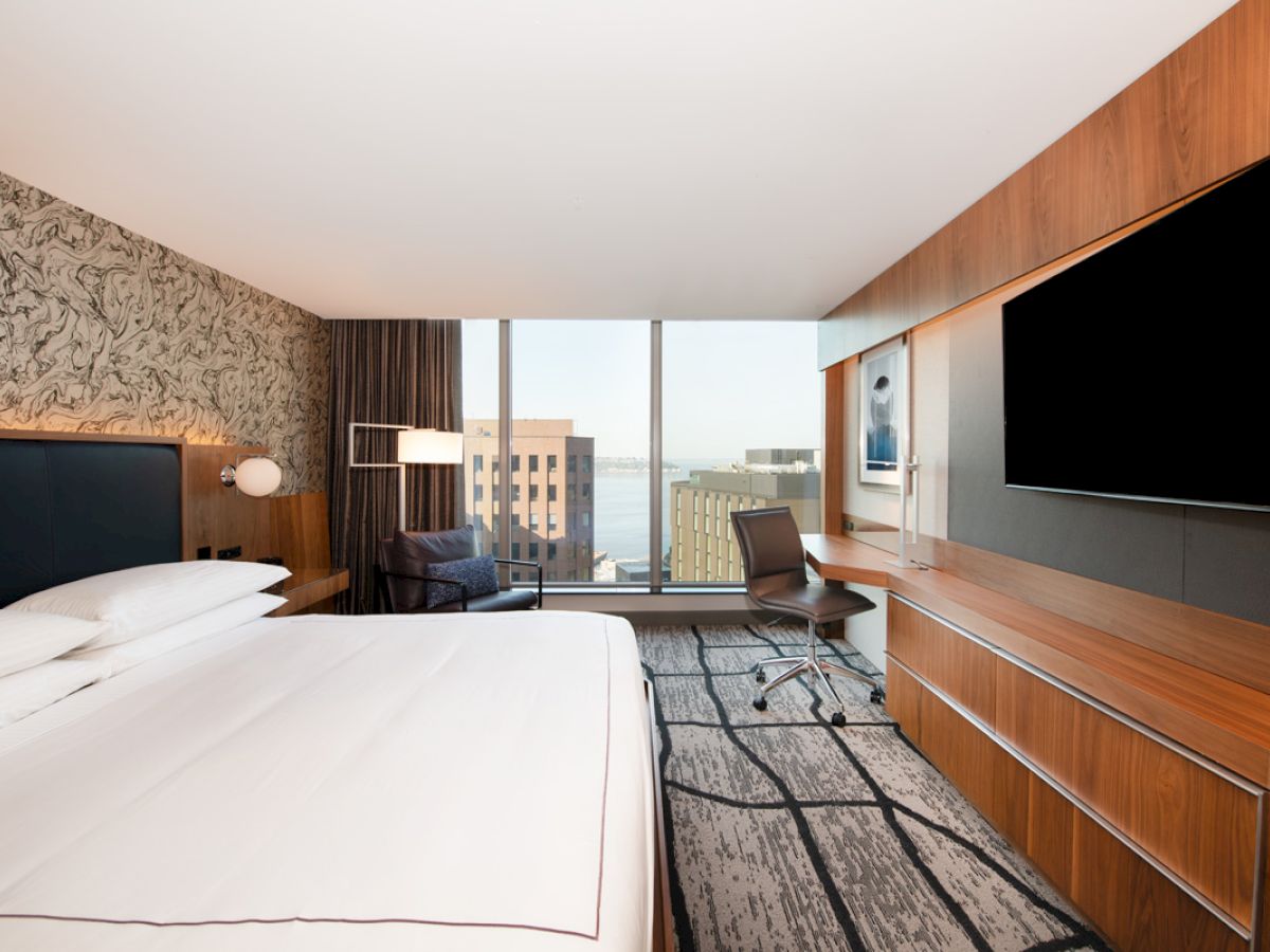 A modern hotel room features a bed, desk, chair, large TV, and floor-to-ceiling windows with a city view.