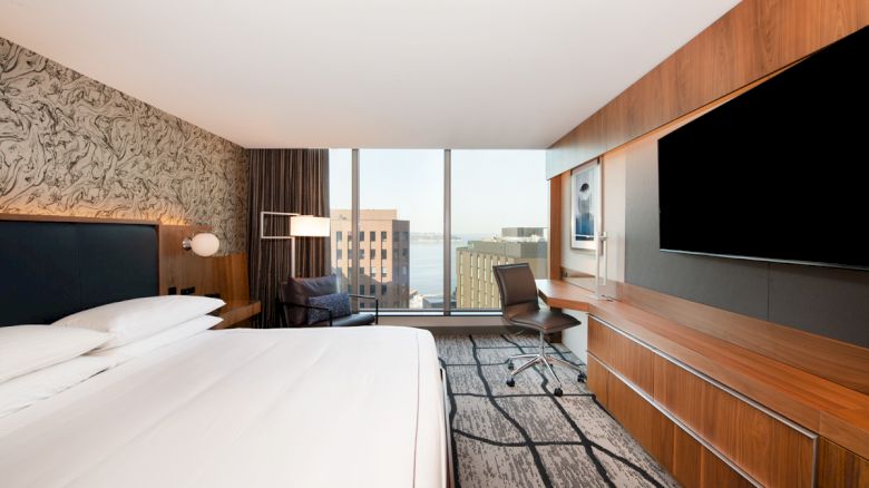 A modern hotel room features a bed, desk, chair, large TV, and floor-to-ceiling windows with a city view.