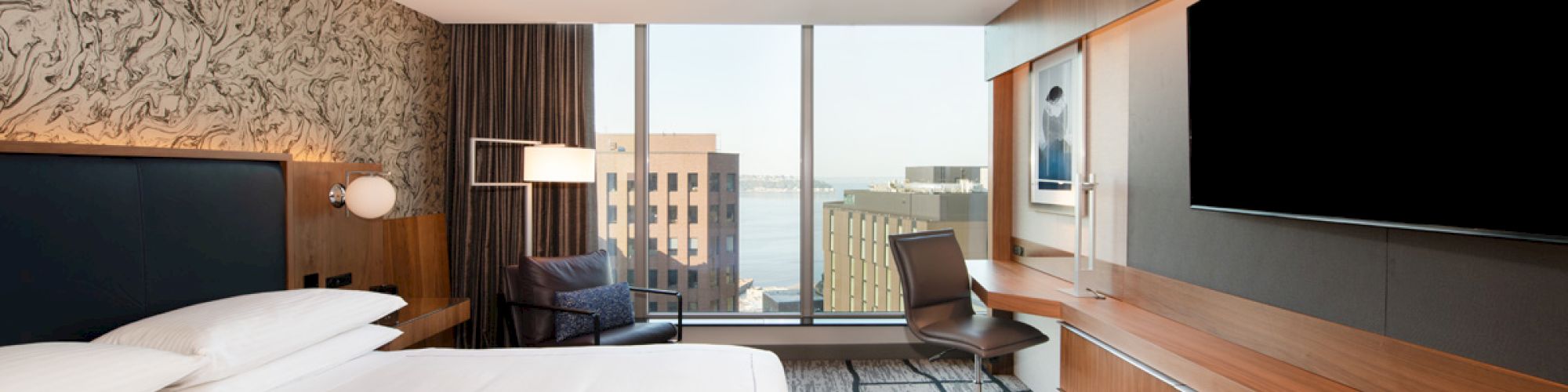 The image shows a modern hotel room with a large bed, desk, chair, wall-mounted TV, and a window view of city buildings and water.